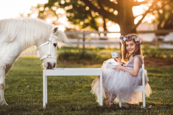little girl wearing big tulle dress and flower crown having a tea party with a white unicorn and smiling at the camera with teacup in hand, photo by Anika Colombo Photography